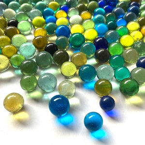May include: A collection of glass marbles in various shades of blue, green, and yellow. The marbles are scattered on a white surface.