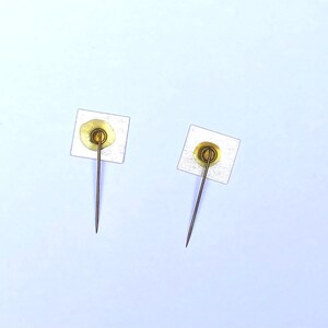 May include: Two gold-colored push pins with white adhesive pads on the back. The pins are on a light blue background.