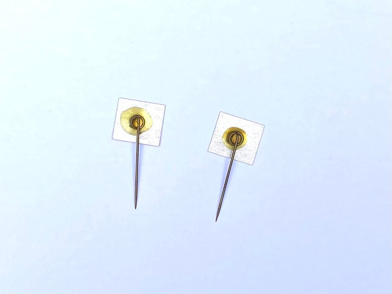 May include: Two gold-colored push pins with white adhesive pads on the back. The pins are on a light blue background.