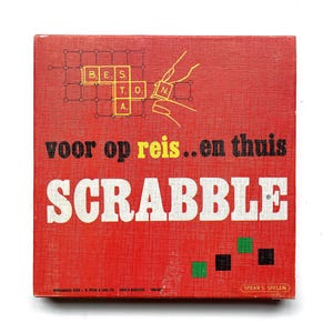 Pode incluir: Caixa vermelha do jogo Scrabble com letras brancas e o texto "voor op reis..en thuis". A caixa apresenta uma grelha com letras e um desenho de mãos a colocar peças. O jogo é da Spear's Spelen.
