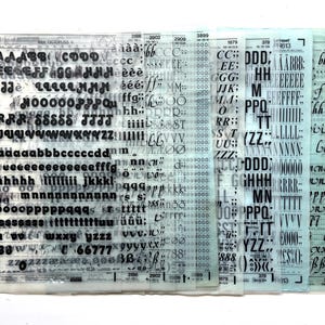Op de afbeelding: Een verzameling transparante vellen met zwarte letters en cijfers. De vellen bevatten verschillende lettertypen en -groottes van letters, cijfers en symbolen. De vellen zijn in een gelaagd formaat gerangschikt, waarbij sommige tekst gedeeltelijk wordt bedekt.
