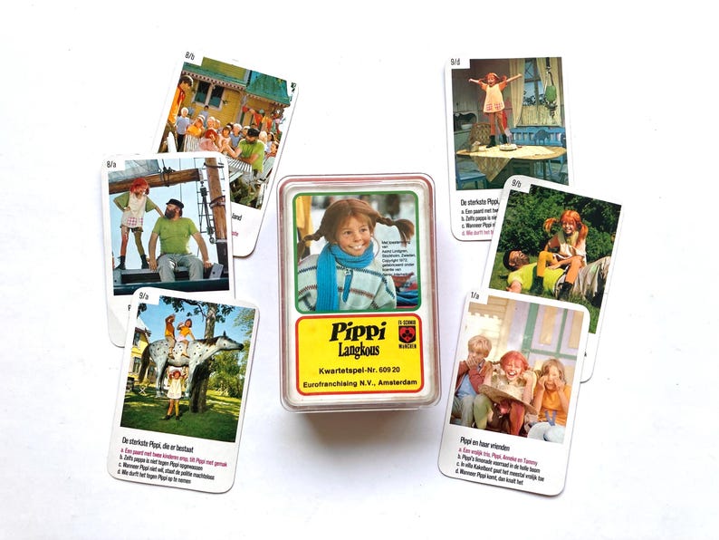 Op de afbeelding: Een vintage Pippi Langkous kaartspel. Het spel bevat een doorzichtige plastic doos met een kaart met Pippi, en verschillende andere kaarten met afbeeldingen van Pippi en haar vrienden. De kaarten zijn wit met kleurrijke illustraties.