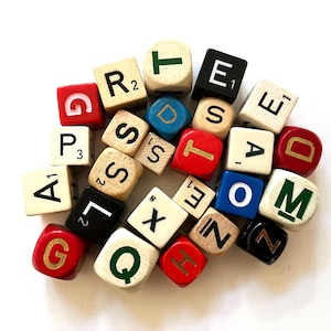 Op de afbeelding: Een verzameling verspreide vierkante houten en plastic spelstenen met diverse letters en numerieke waarden. De stenen zijn gekleurd in rood, blauw, zwart en natuurlijke houttinten, met bedrukte tekens in contrasterende kleuren zoals wit, goud en groen.