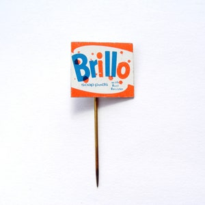 May include: A vintage Brillo soap pads pin with a red and white background. The pin features the Brillo logo in blue and the text "soap pads with Rust Resistor".