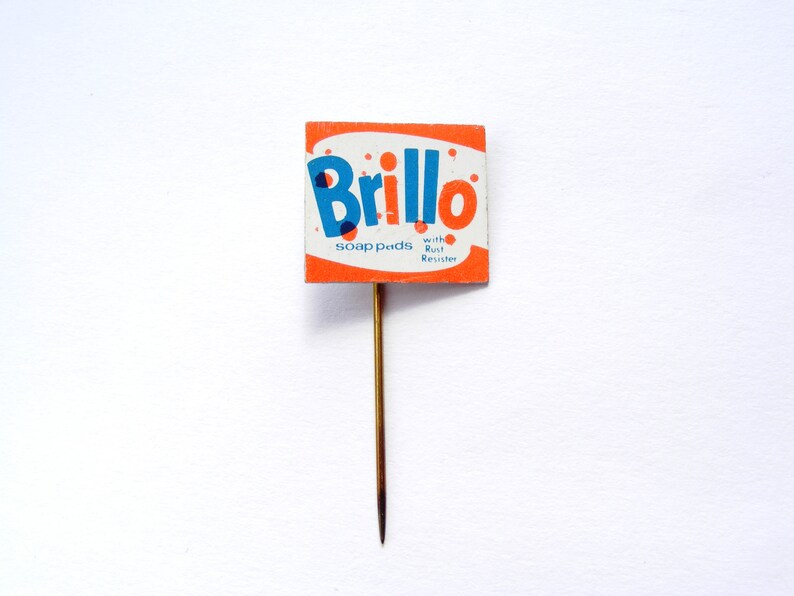 May include: A vintage Brillo soap pads pin with a red and white background. The pin features the Brillo logo in blue and the text "soap pads with Rust Resistor".