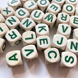Op de afbeelding: Een verzameling houten letterdobbelstenen met groene letters. De dobbelstenen zijn verspreid over een wit oppervlak en tonen verschillende letters. De dobbelstenen zijn lichtbeige van kleur en de groene letters zijn duidelijk zichtbaar.