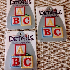 May include: Three identical packages of iron-on applique patches, each featuring a stack of three square blocks with the letters A, B, and C. The packaging is shaped like an iron and displays the text IRON-ON DETAILS Quick, Easy Applique Fun!