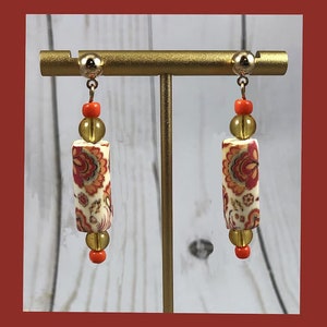 May include: A pair of dangle earrings featuring a white cylinder bead with a red and orange floral pattern, a gold bead, and a small orange bead. The earrings are displayed on a gold stand.