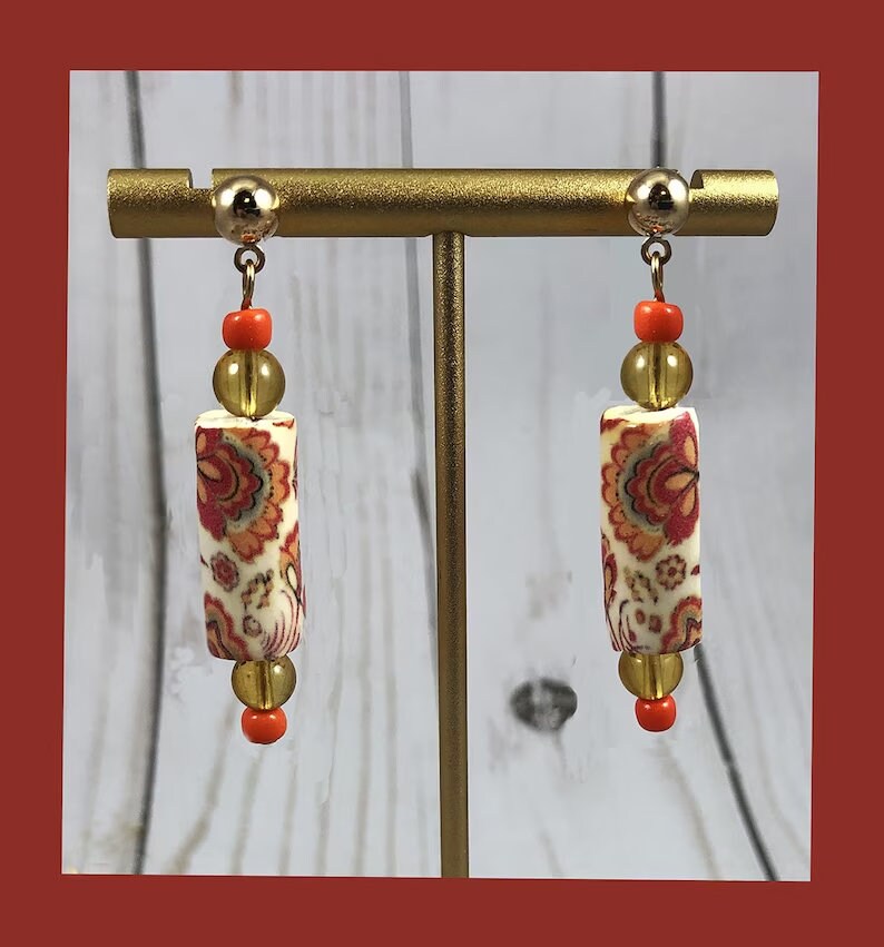 May include: A pair of dangle earrings featuring a white cylinder bead with a red and orange floral pattern, a gold bead, and a small orange bead. The earrings are displayed on a gold stand.