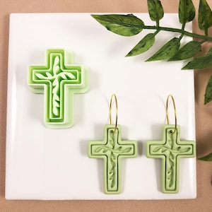 May include: A pair of green cross-shaped earrings with gold hoops. The earrings are made of polymer clay and have a detailed design.