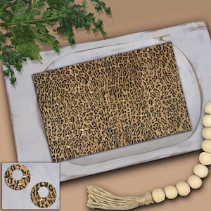 May include: A rectangular cork board with a leopard print pattern and gold accents. The board is displayed on a white tray with a decorative wooden bead garland and a sprig of greenery. A pair of matching earrings is in the lower left corner.