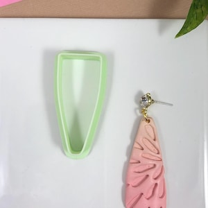 May include: A pink and white polymer clay earring with a leaf design. The earring is shown next to a green silicone mold.