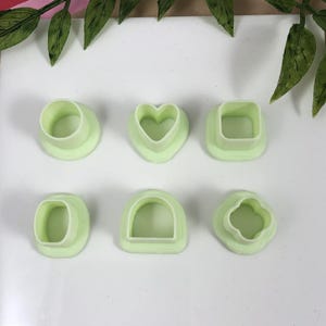 May include: Six light green polymer clay cutters in various shapes: a circle, a heart, a square, an oval, a rounded arch, and a four-leaf clover. These tools are likely used for crafting or baking.
