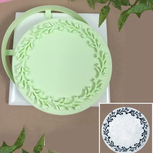May include: A light green, circular silicone mold with a floral border design. The mold is sitting on a white square surface. A smaller image shows a finished plate with a similar design.