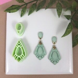 May include: Two pairs of green dangle earrings with gold accents. The earrings are displayed on a white surface with two silicone molds for making the earrings.