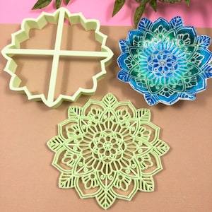 May include: Three decorative items: a white plastic cookie cutter in the shape of a four-pointed star, a green and blue mandala-patterned ceramic dish, and a green plastic mandala-shaped cookie cutter.