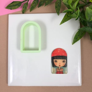 May include: A white ceramic tile with a green plastic arch-shaped mold and a resin charm of a girl with black hair wearing a red hat and a red coat.