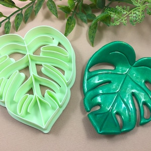 May include: Two monstera leaf shapes, one is a green plastic cookie cutter and the other is a green plastic leaf.