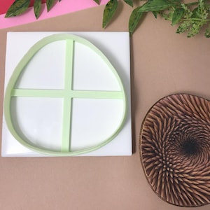 May include: A light green plastic egg-shaped cookie cutter with a cross-shaped support sits on a white square. A wooden, spiral-carved object is next to it. The background is a mix of pink, brown, and green.