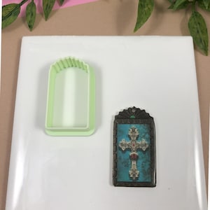 May include: A green plastic cookie cutter shaped like a rectangle with a scalloped top edge. Next to it is a small framed image of a cross with a blue background and ornate details.