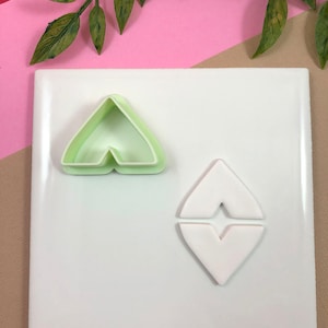 May include: A light green plastic cookie cutter in the shape of a stylized heart, next to a white heart-shaped fondant cutout. The cutter is on a white surface with a pink and tan background, with green leaves in the top corners.