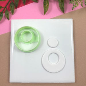 May include: A set of three white plastic cookie cutters. The largest cutter is a ring with a smaller circle inside. The medium cutter is a circle. The smallest cutter is a small circle.