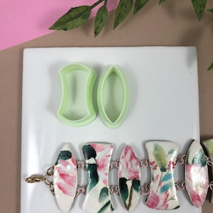 May include: A white ceramic bracelet with a floral pattern and gold chain links. The bracelet is made of individual pieces that are connected together. Two green plastic cookie cutters are shown in the background.