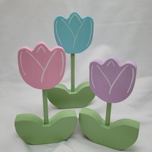 May include: Three wooden tulips with green leaves. The tulips are painted in pastel colors: pink, blue, and purple.