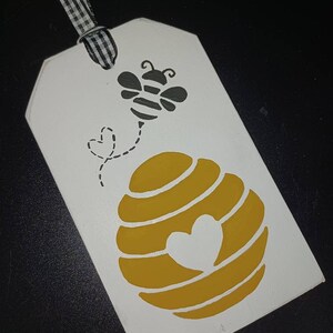 May include: A white wooden tag with a black and white gingham ribbon. The tag features a yellow beehive with a heart cutout and a black and yellow bee with a dotted trail. A charming, decorative item.