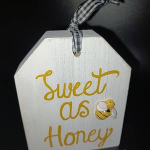 May include: White wooden sign with a black and white checkered ribbon attached. The sign says "Sweet as Honey" in yellow lettering and has a small yellow and black bee attached.