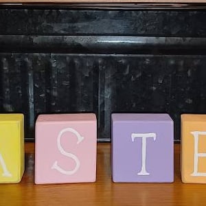 May include: Wooden blocks spelling out the word "EASTER" in pastel colors. The blocks are arranged in a row on a wooden surface. Two white wooden bunny figurines are on either side of the blocks.
