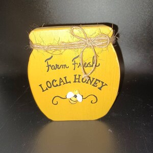 May include: A yellow wooden honey jar with a twine bow and a painted bee. The jar says "Farm Fresh Local Honey".