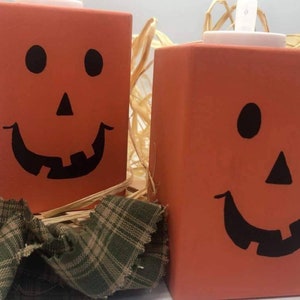 May include: Two orange wooden pumpkin-shaped boxes with black painted jack-o'-lantern faces. The boxes are sitting on a bed of straw and a green and brown plaid fabric.