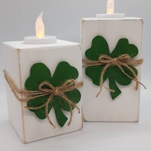 May include: Two white wooden candle holders with a green shamrock design and a brown twine bow. Each holder has a small white LED tea light inside.