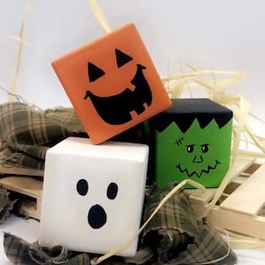 May include: Three wooden blocks painted with Halloween characters. The first block is orange and has a jack-o'-lantern face. The second block is green and has a Frankenstein face. The third block is white and has a ghost face.