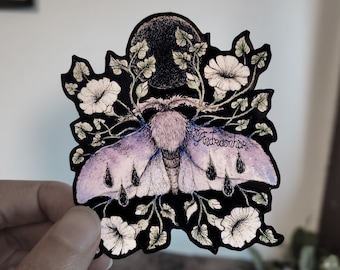 May include: A black and white illustration of a moth with purple wings and black teardrop-shaped markings. The moth is surrounded by white flowers and green leaves. The artist's signature, "Andreaschor", is visible in the lower right corner.