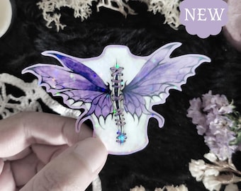 Puede incluir: Una pegatina de mariposa iridiscente púrpura y azul con una costura cosida en el centro. La pegatina está sobre un fondo blanco y tiene un acabado holográfico.