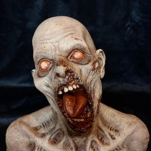 Puede incluir: Busto de zombi detallado con piel pálida y ojos naranja brillante. La boca está abierta, mostrando dientes y una lengua roja. El efecto general es grotesco e inquietante, con detalles realistas.