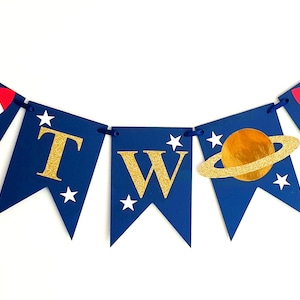 May include: A blue and gold banner with the word "TWO" in gold glitter letters. The banner is decorated with stars, a rocket ship, and a planet with rings.