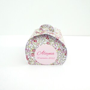 Pu&ograve; includere: Una piccola scatola regalo bianca con un motivo floreale rosa e giallo. La scatola ha un cerchio rosa con il nome "Aleyna" e la data "16 settembre 2022" scritta in nero.