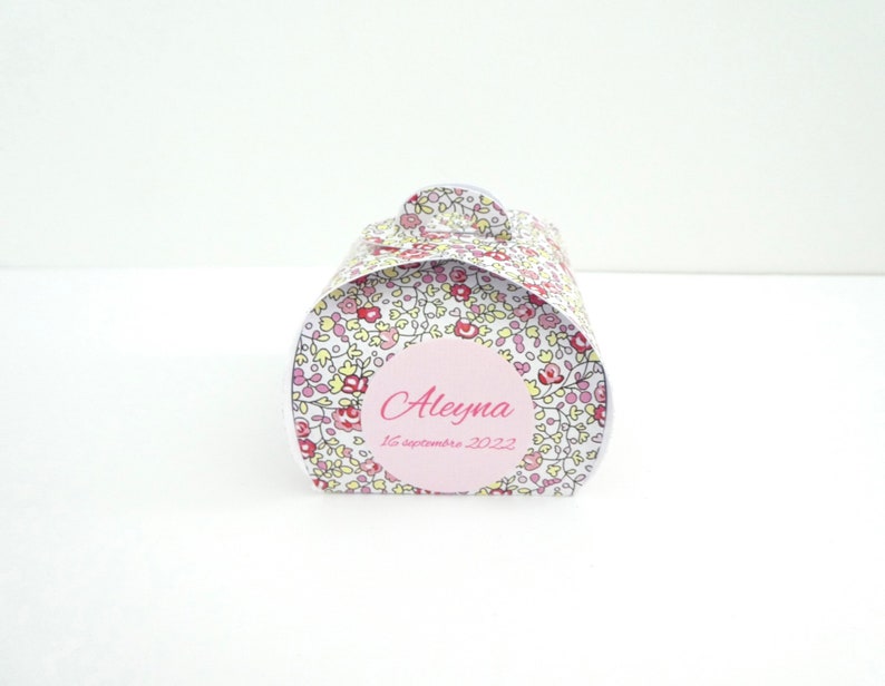 Pu&ograve; includere: Una piccola scatola regalo bianca con un motivo floreale rosa e giallo. La scatola ha un cerchio rosa con il nome "Aleyna" e la data "16 settembre 2022" scritta in nero.
