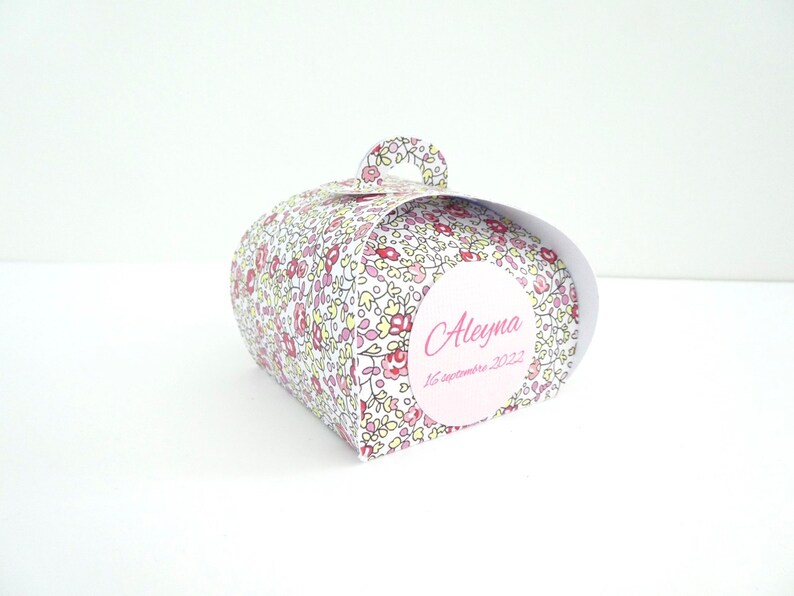 Pu&ograve; includere: Una scatola regalo bianca con un motivo floreale in tonalit&agrave; rosa, giallo e rosso. La scatola ha un manico e un'etichetta rotonda con il nome "Aleyna" e la data "16 settembre 2022" scritta in rosa.