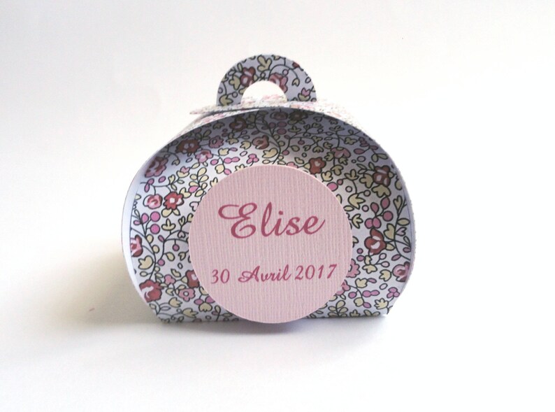 Pu&ograve; includere: Una piccola scatola regalo bianca con motivo floreale e un'etichetta circolare rosa che dice "Elise 30 Avril 2017".