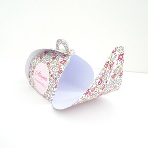 Pu&ograve; includere: Una scatola regalo di carta bianca a forma di teiera con un motivo floreale rosa, giallo e verde. La scatola ha un cerchio rosa con il nome "Aleyma" e la data "settembre 2022" stampata su di essa.