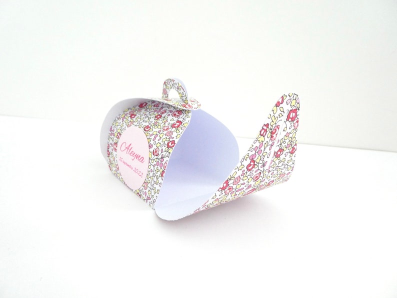 Pu&ograve; includere: Una scatola regalo di carta bianca a forma di teiera con un motivo floreale rosa, giallo e verde. La scatola ha un cerchio rosa con il nome "Aleyma" e la data "settembre 2022" stampata su di essa.