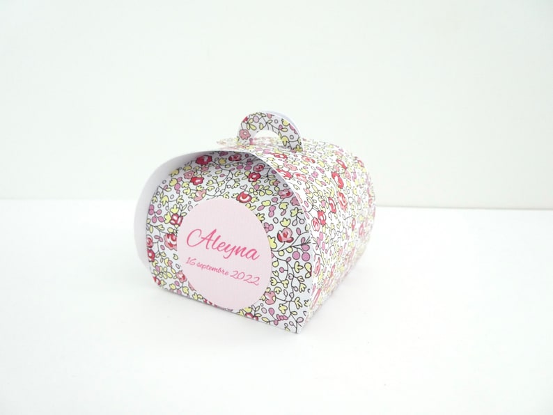 Pu&ograve; includere: Una piccola scatola regalo bianca con un motivo floreale e un'etichetta circolare rosa che dice "Aleyna 16 settembre 2022".