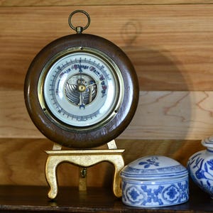 May include: A collection of antique decorative items on a wooden shelf. Includes a wooden barometer on a gold stand, two blue and white porcelain containers, a brass lantern, and two vintage books with blue covers.