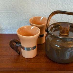 May include: A brown enamel teapot with a wooden knob on the lid, and two peach-colored ceramic mugs with wooden handles and silver bands.