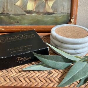 Pu&ograve; includere: Un set di quattro sottobicchieri in marmo bianco con inserti in sughero. I sottobicchieri sono confezionati in una scatola nera con il testo "The Irish View Collection" e "Quality Coasters by John Hinde Limited".