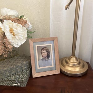 May include: A rectangular light wood picture frame containing a vintage portrait of a person with shoulder-length hair wearing a light blue collared shirt. The frame features a light gray matting and is positioned next to a gold-toned metal lamp.